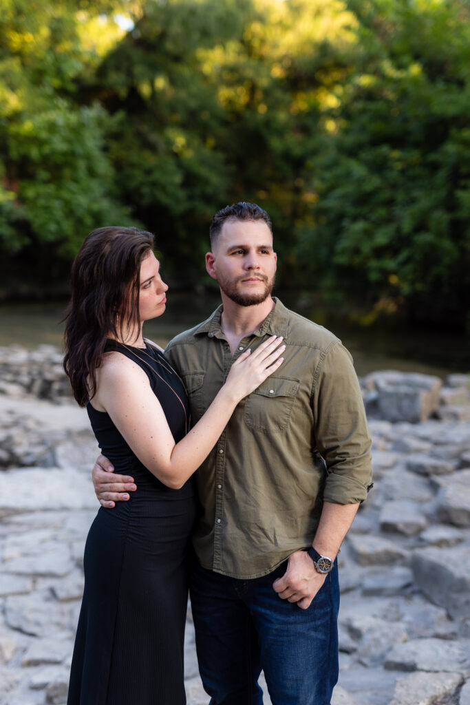 Engaged woman looking at fiancé with hand on his chest surrounded by stones and trees during Richardson Park engagement session in TX