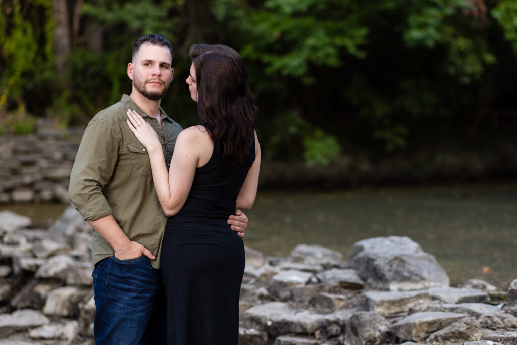 Engaged woman with hand on fiancé's chest while he looks into camera surrounded by stones and trees during Richardson Park engagement session in TX