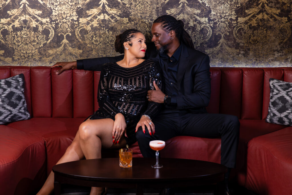 Man and woman sitting on red couch with gold wallpaper during anniversary session at Thompsons Bookstore Speakeasy in downtown FW