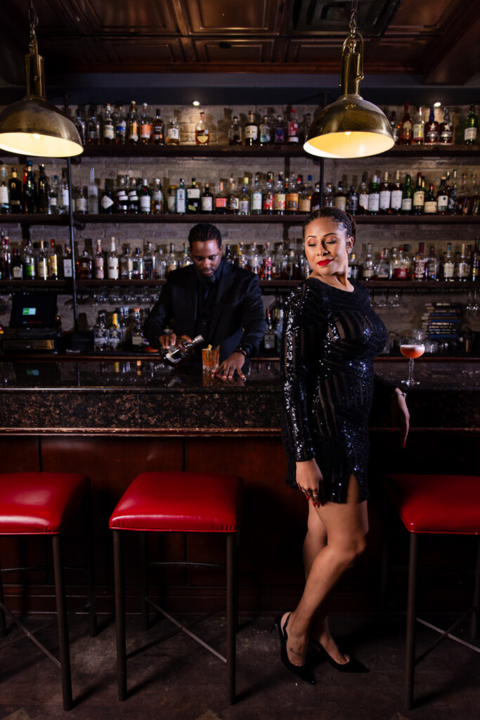 Woman standing in front of bar wearing black dress looking over her shoulder while man makes cocktail behind bar at Thompsons Bookstore and Speakeasy during anniversary portrait session