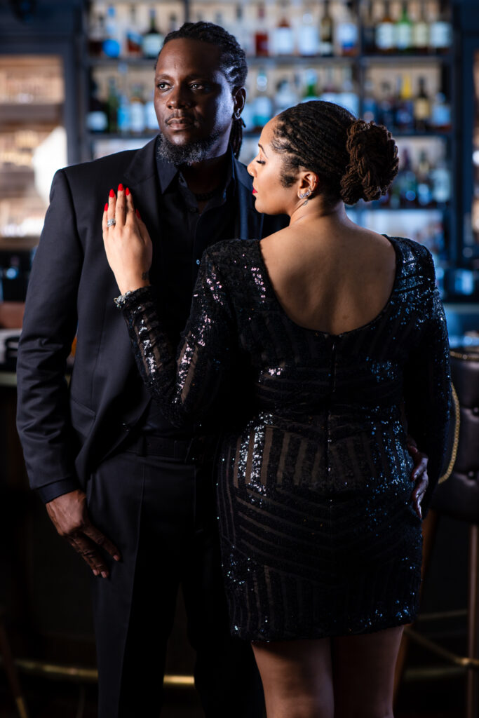 Woman leaning against man's chest in all black during chic anniversary portrait session at Thompsons Bookstore Speakeasy in downtown Fort Worth