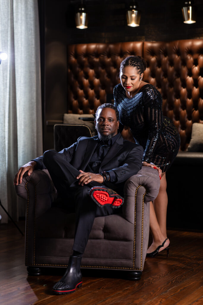 Man sitting in chair revealing red bottom boots while woman leans behind in all black during anniversary photo session in vintage Fort Worth speakeasy