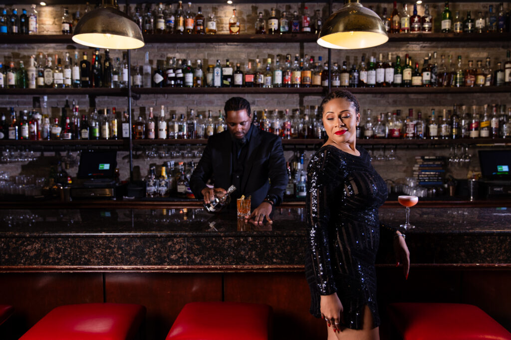 Woman standing in front of bar wearing black dress looking over her shoulder while man makes cocktail behind bar at Thompsons Bookstore and Speakeasy in downtown Fort Worth during portrait session