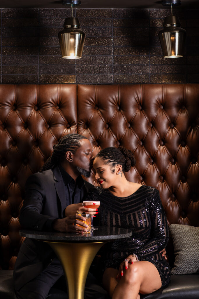 Man kissing woman's temple while sitting together at a cocktail table at a speakeasy