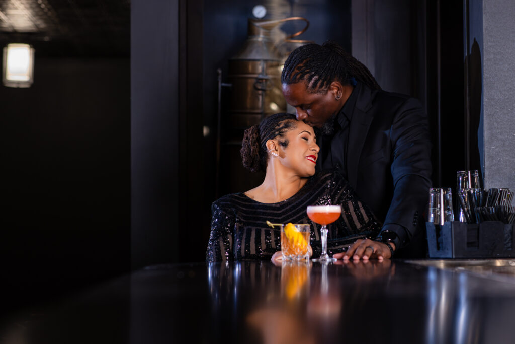 Man kissing woman who is sitting at the bar on the top of her head with cocktails in front of them