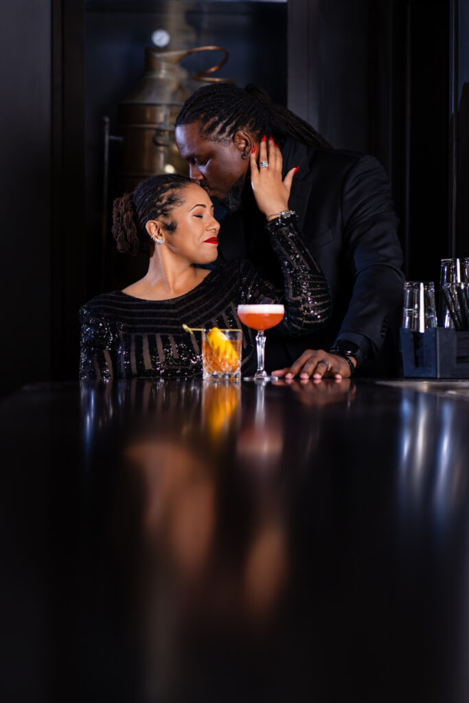 Man kissing woman on the top of head while she sits at a bar with cocktails in front of them during portrait session