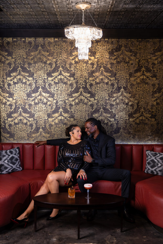 Man and woman sitting on red couch with gold wallpaper during unique anniversary session at Thompsons Bookstore Speakeasy in downtown Fort Worth