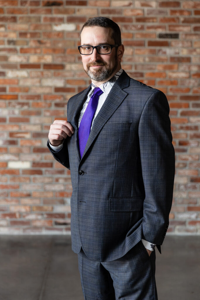 groom in dark grey ted baker suit and purple tie standing with hand in pocket and holding onto lapel in front of brick wall