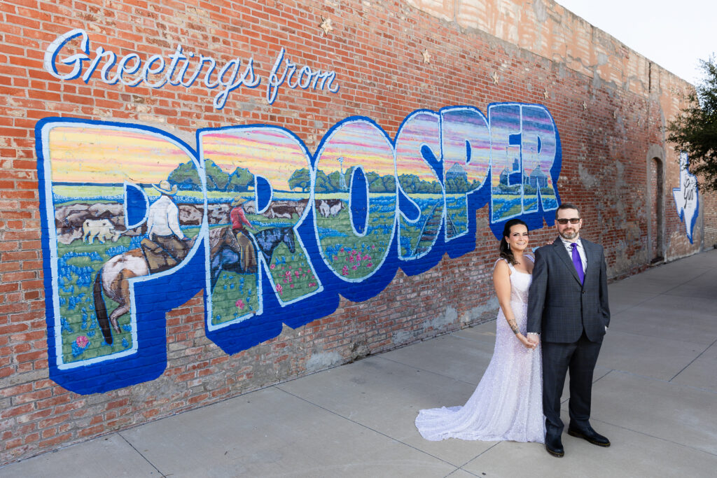 bride in a Galia Lahav Oriana sparkling a-line wedding dress standing with groom in a dark grey ted baker suit in front of Greetings from Prosper mural sign in downtown Prosper TX