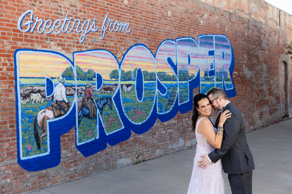 bride in a Galia Lahav Oriana sparkling a-line wedding dress standing with groom in a dark grey ted baker suit in front of Greetings from Prosper mural sign in downtown Prosper Texas