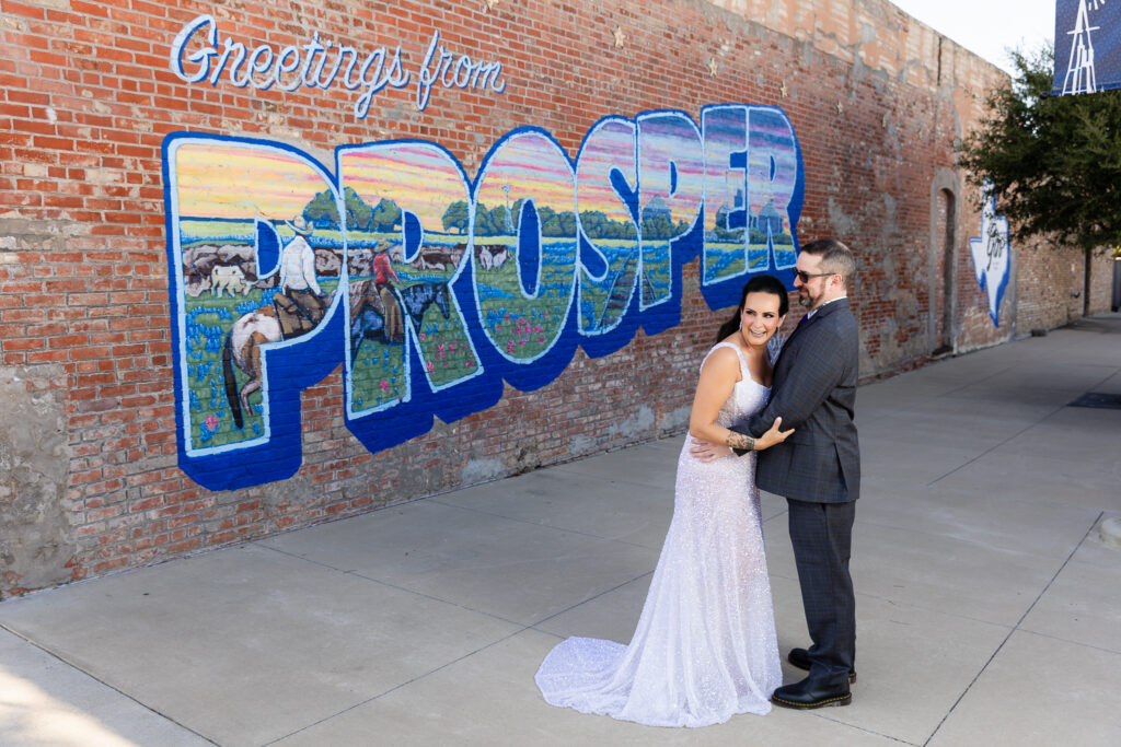 bride in a Galia Lahav Oriana sparkling a-line wedding dress laughing with groom in a dark grey ted baker suit in front of Greetings from Prosper mural sign in downtown Prosper TX