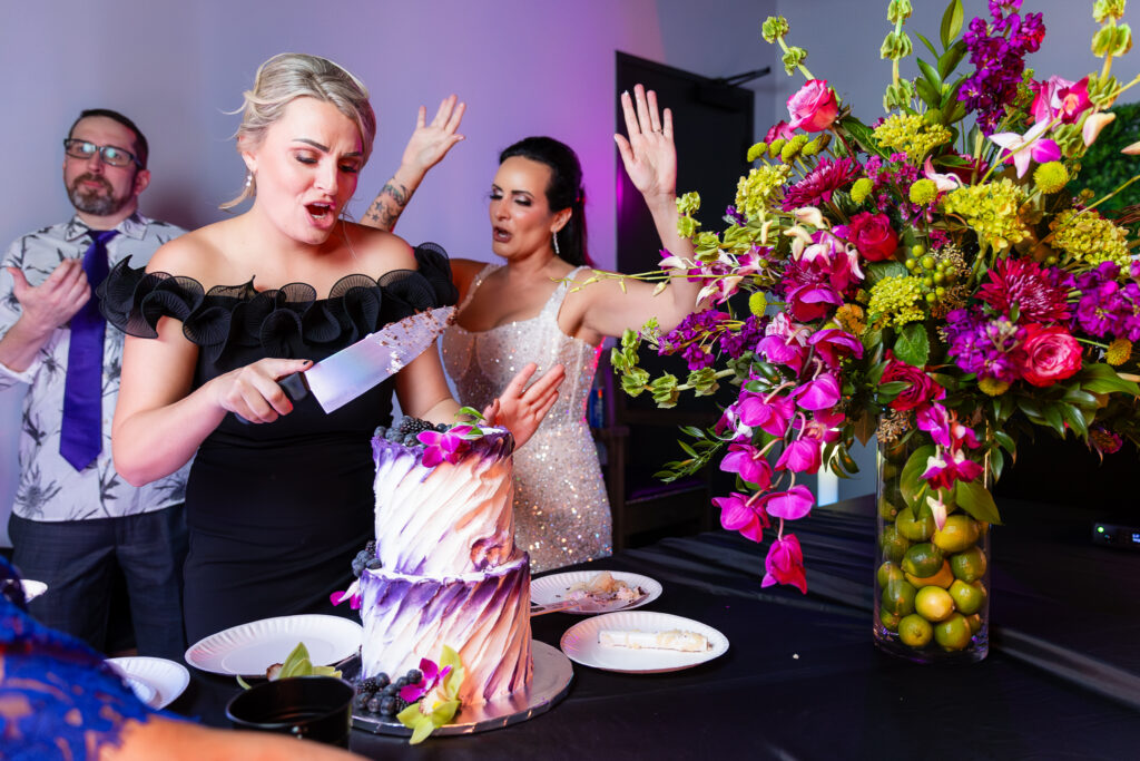 maid of honor cutting 2-tier white and purple The London Baker cake while bride dances behind