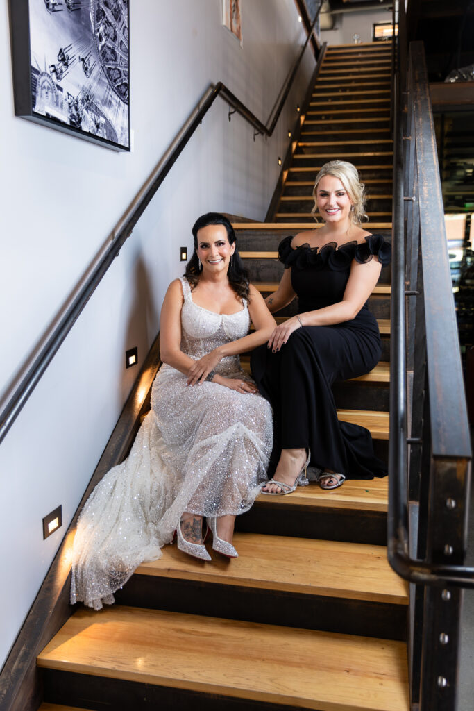 bride in a Galia Lahav Oriana sparkling a-line wedding dress sitting on staircase at Prosper Wine House with maid of honor in black dress smiling at camera