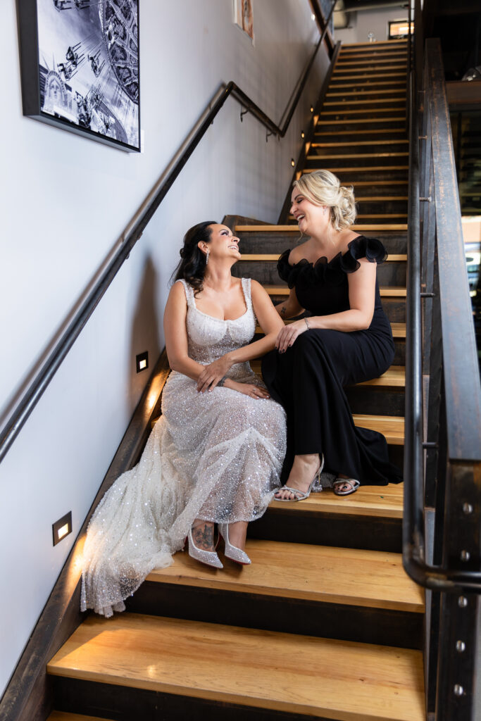 bride in a Galia Lahav Oriana sparkling a-line wedding dress sitting on staircase at Prosper Wine House with maid of honor in black dress smiling at each other