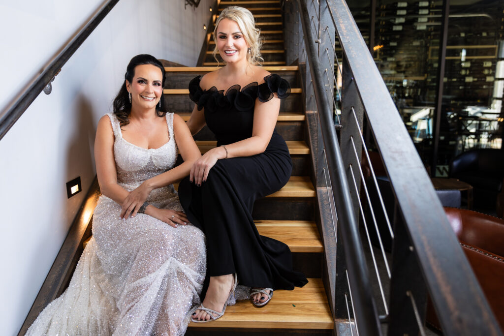 bride in a Galia Lahav Oriana sparkling a-line wedding dress sitting on staircase at Prosper Wine House with maid of honor in black dress smiling