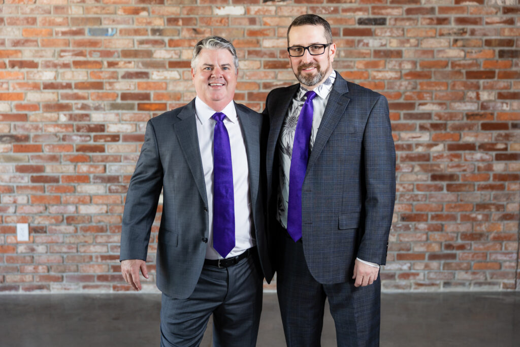 groom and best man standing together in dark grey ted baker suits in front of brick wall