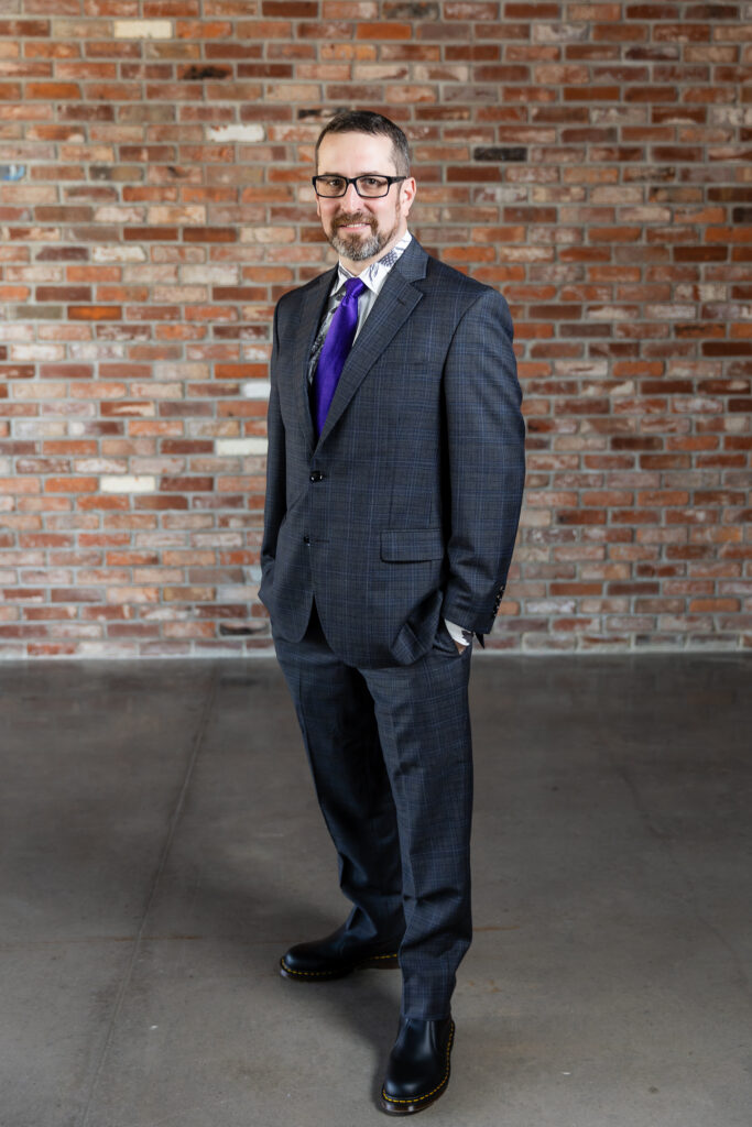 groom in dark grey ted baker suit and purple tie with hands in pockets in front of brick wall