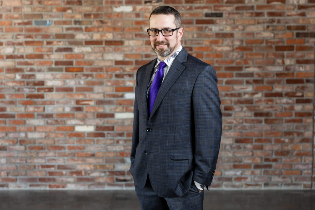 groom standing in dark grey ted baker suit and purple tie with hands in pockets in front of brick wall