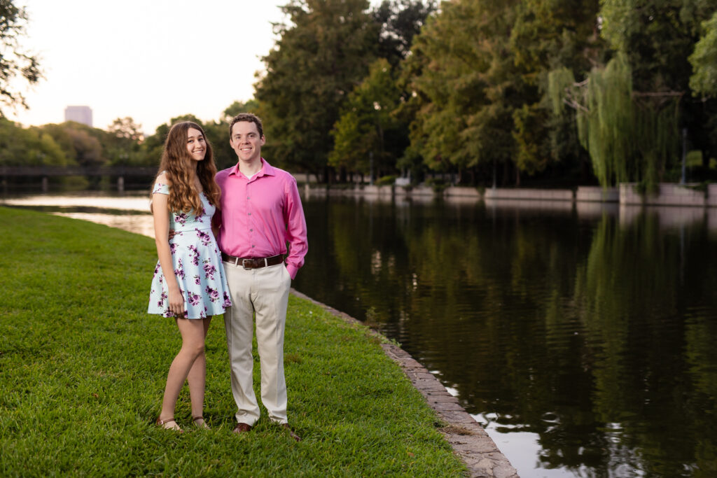 engaged couple in blue and pink dress and pink shirt standing near lake at sunrise during lakeside park engagement session