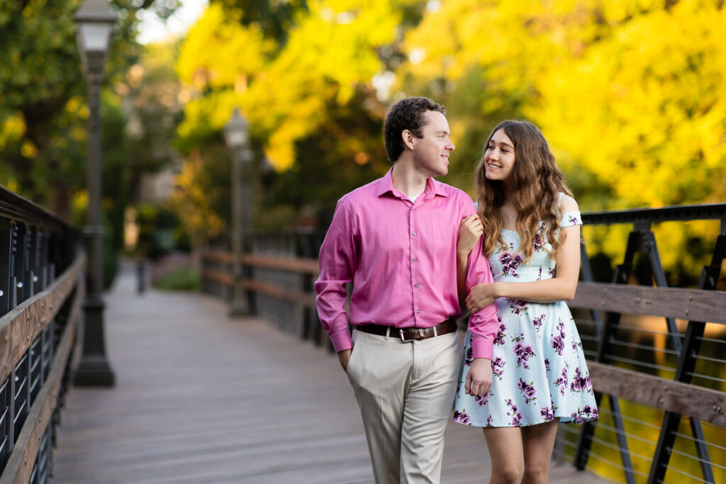 engaged couple walking arms interlocked across wooden bridge with lampposts while smiling at each other during sunrise lakeside park engagement session in Dallas