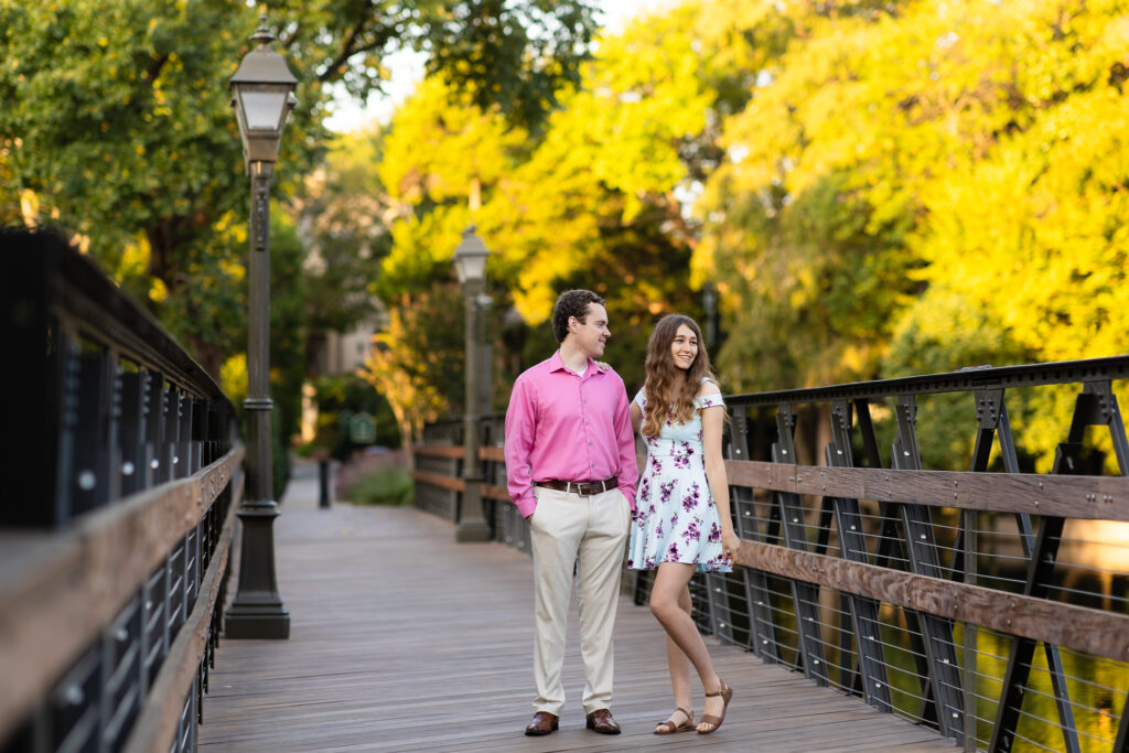 girl in blue floral dress and man in pink shirt standing on wooden bridge together smiling out at trees and lake during dallas engagement session at lakeside park