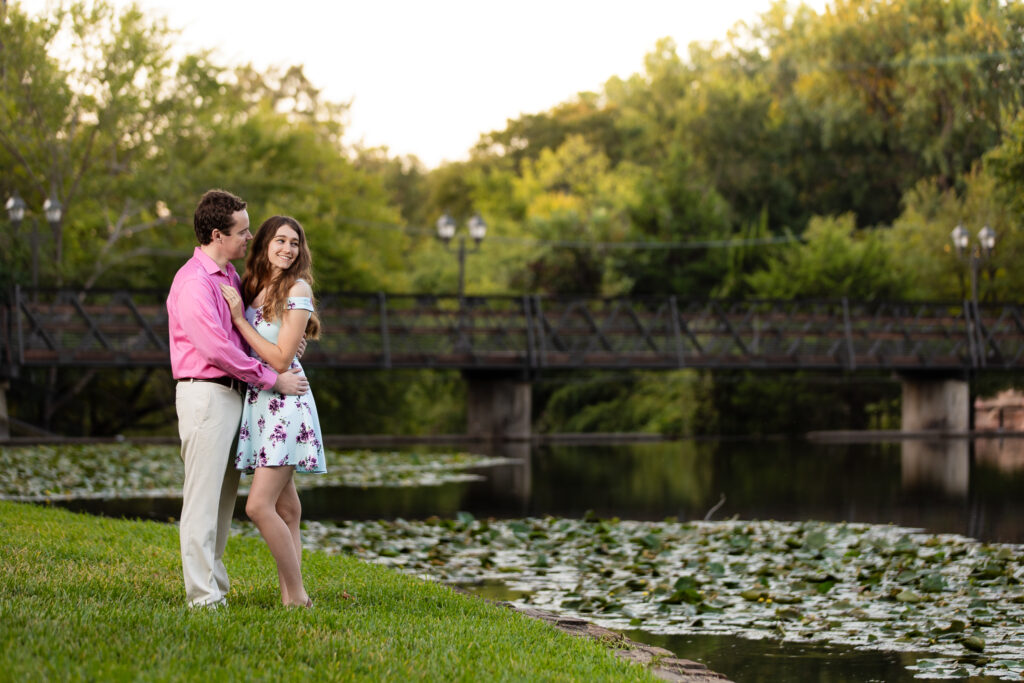 man wearing pink shirt whispering in fiancée's ear wearing blue dress and laughing towards lake and wooden bridge at lakeside park in Dallas