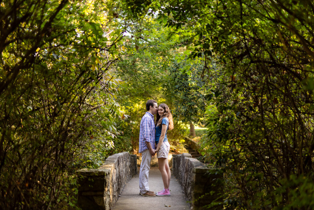 man kissing woman on cheek while holding hands on stone bridge surrounded by trees at Prather Park