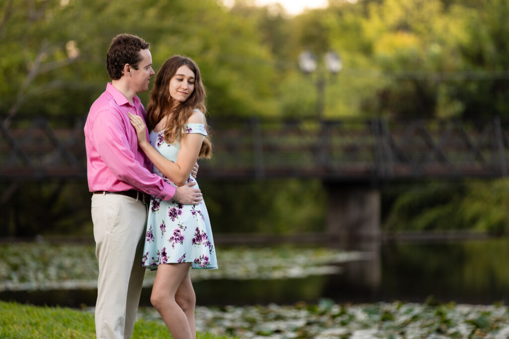 engaged couple embracing by lake in front of wooden bridge at sunrise at lakeside park engagement session