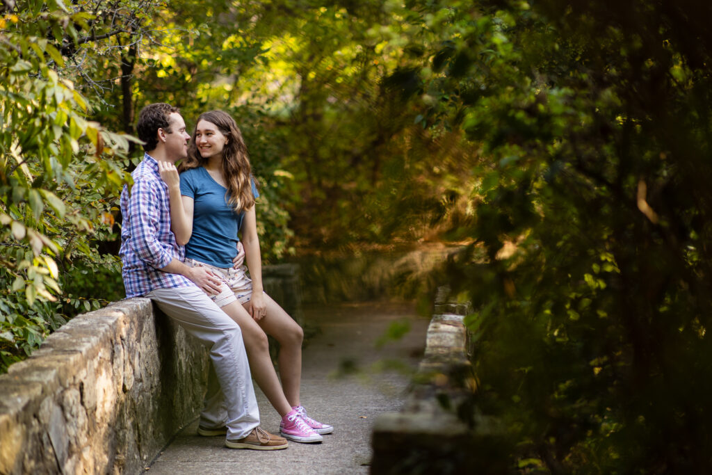 engaged couple sitting on stone bridge wall smiling at each other during engagement session at Prather Park