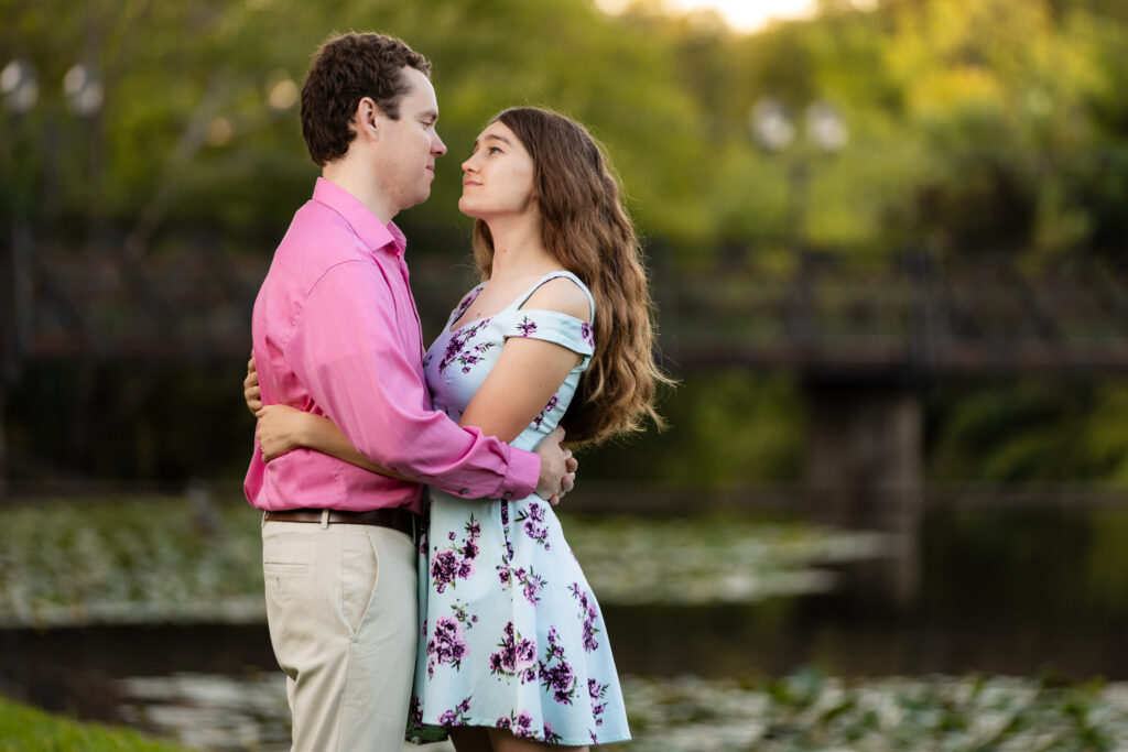 engaged couple looking into each other's eyes while embracing by lake and wooden bridge during lakeside park engagement session