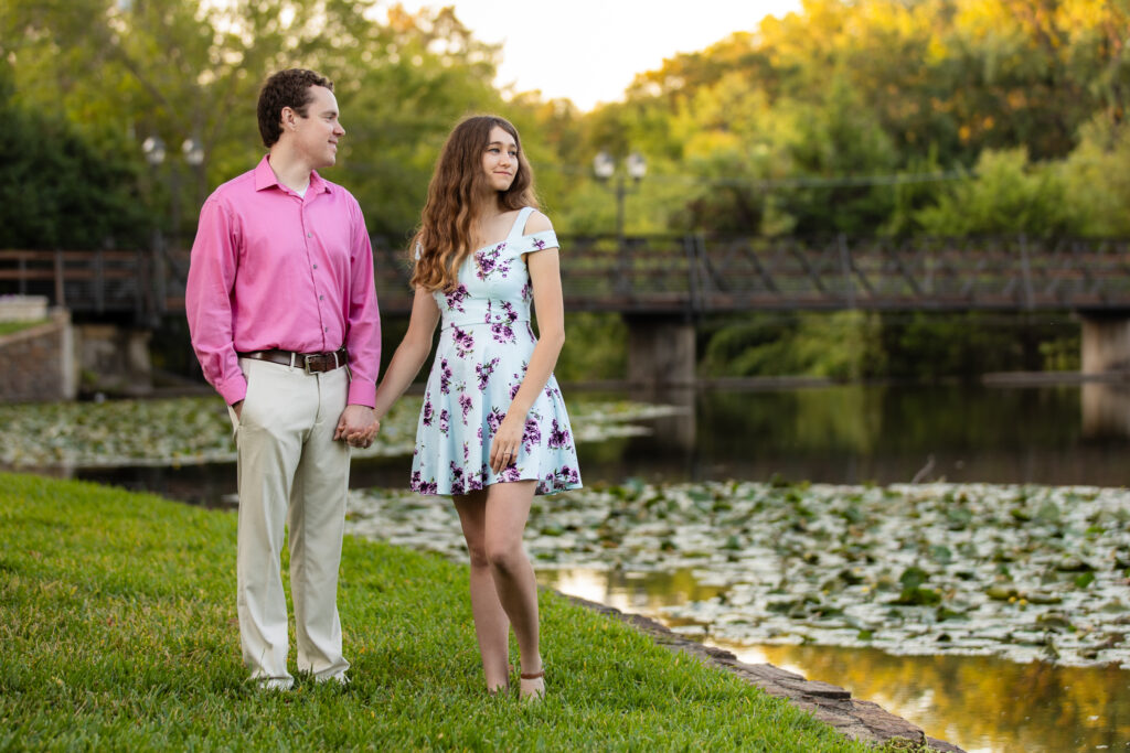 engaged couple standing hand in hand while looking out at lake at sunrise during lakeside park engagement session in Dallas Texas