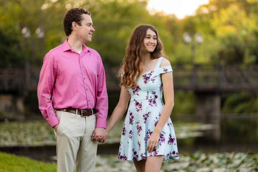 man in pink smiling at woman wearing blue dress with pink and purple florals by the lake at lakeside park engagement session