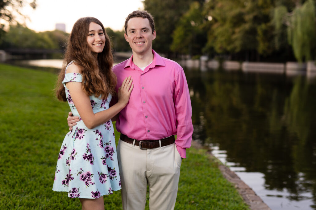 girl in blue dress with pink and purple flowers standing with hand on chest of fiancé wearing pink shirt by lake during  lakeside park engagement session