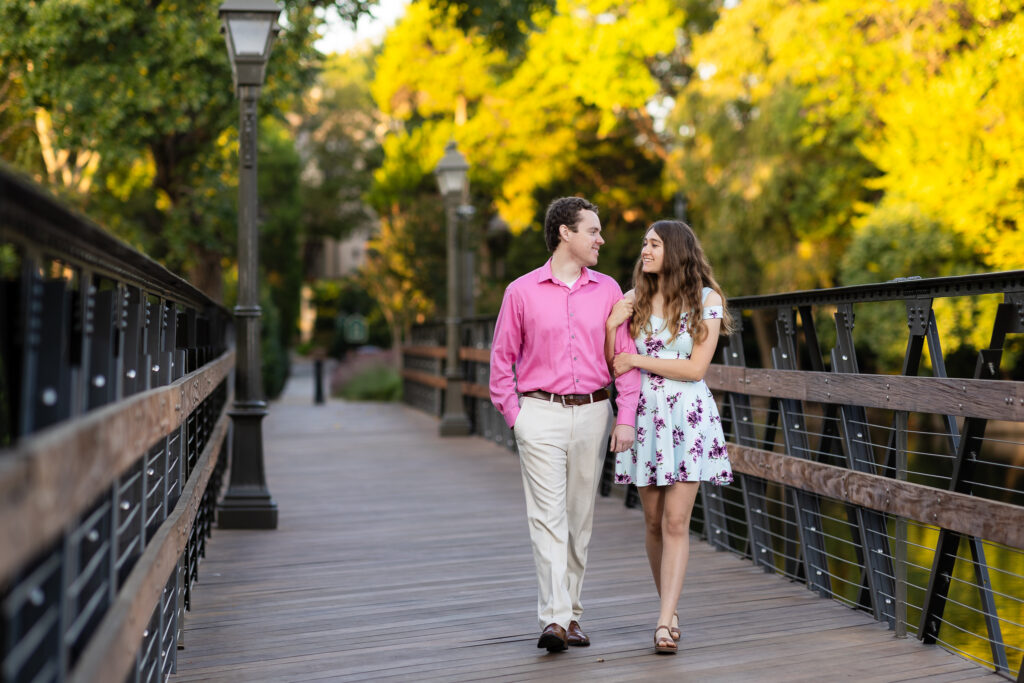 engaged couple walking arms interlocked across wooden bridge with lampposts during sunrise lakeside park engagement session