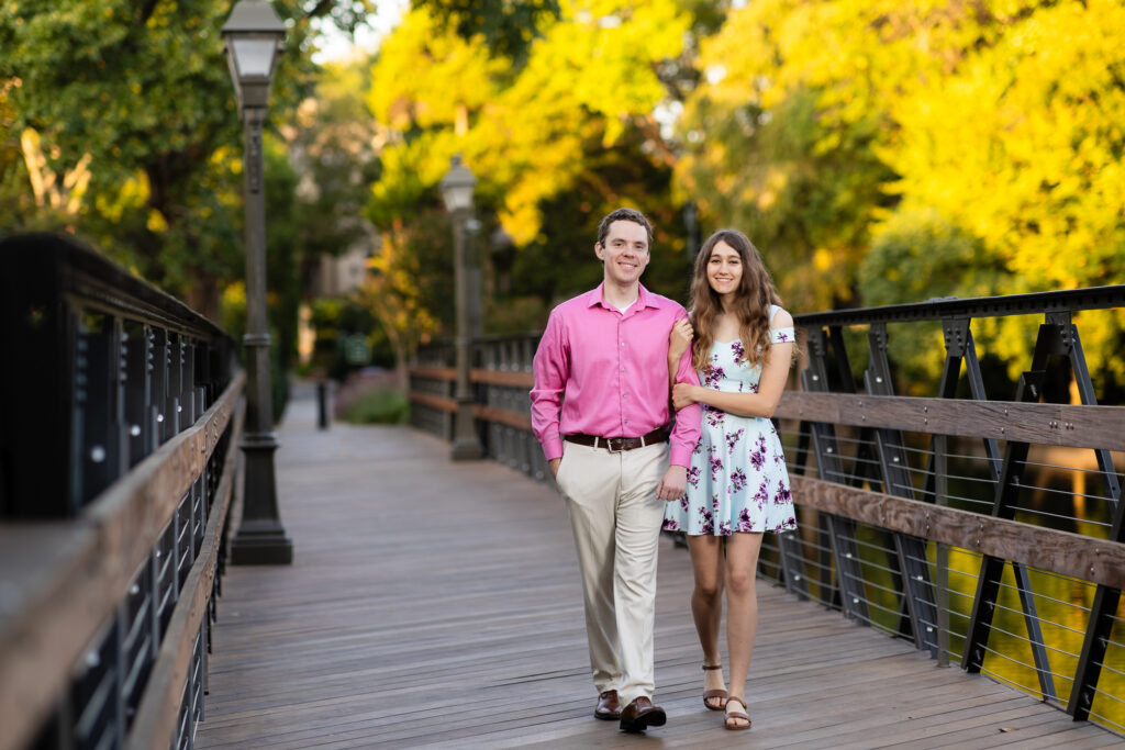 engaged couple walking arms interlocked across wooden bridge with lampposts while smiling during sunrise lakeside park engagement session