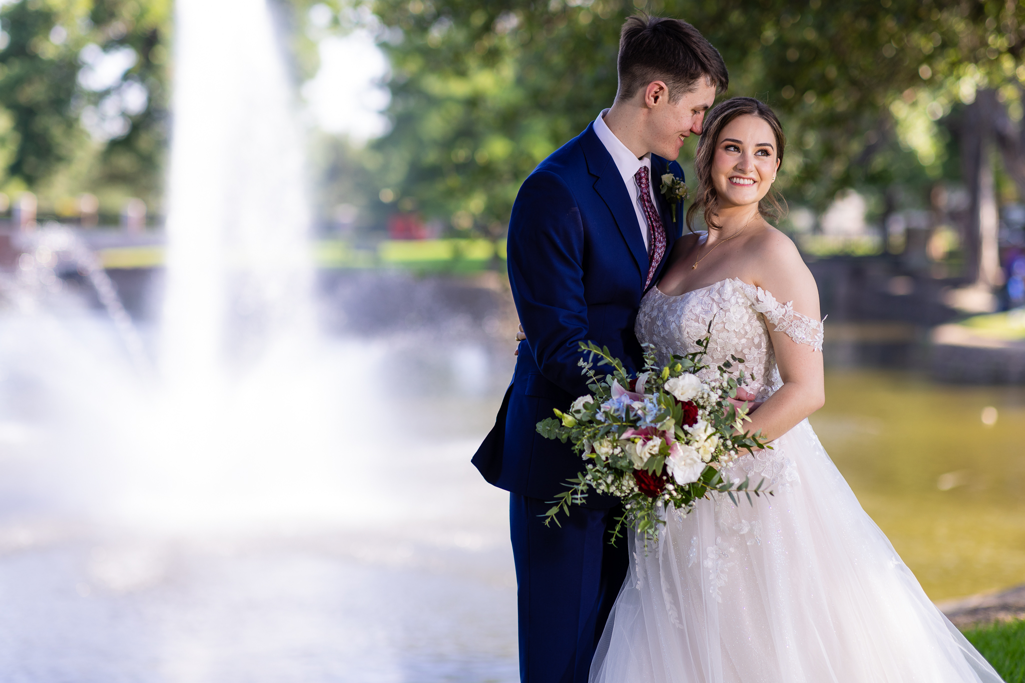 groom in navy blue suit and red tie smiling at bride who's smiling off the side while in an off the shoulder lace wedding dress and holding a blue, red, white and green bridal bouquet in front of huge water fountain feature