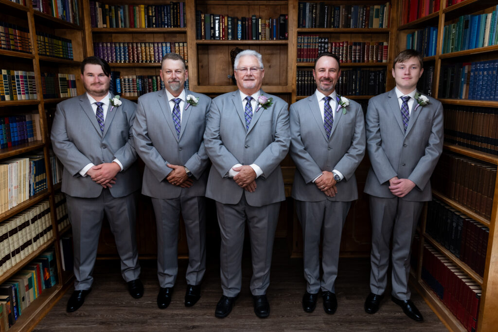 groom with groomsmen in grey suits standing in line smiling at the camera inside The Abbey's groom's suite library