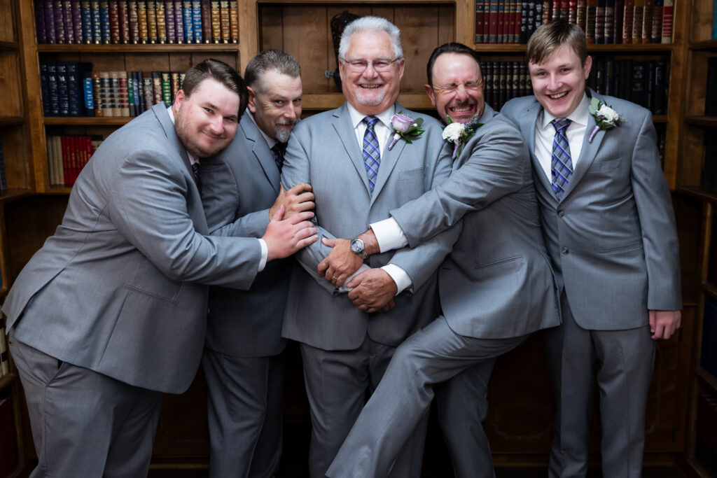 groomsmen hugging in on groom laughing all wearing grey suits inside The Abbey's groom's suite in Justin