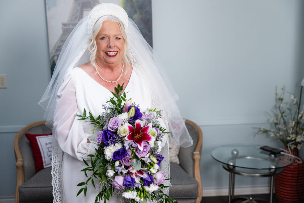 bride holding purple, white and red bouquet wearing a white dress and a veil handmade in 1974