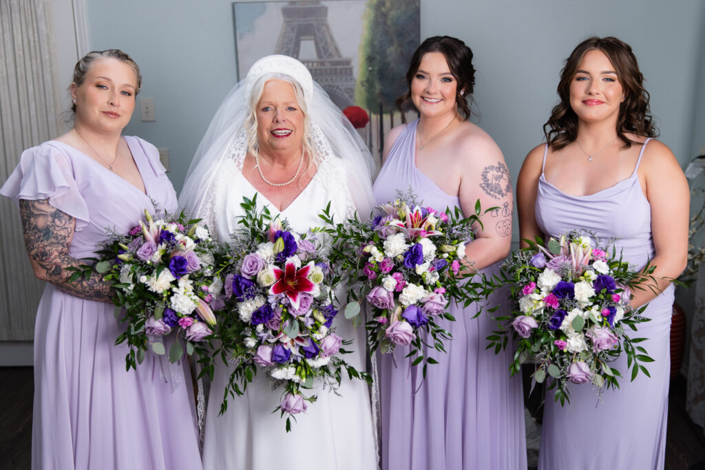 elderly bride smiling with bridemaids holding colorful purple bouquets in bridal suite at The Abbey