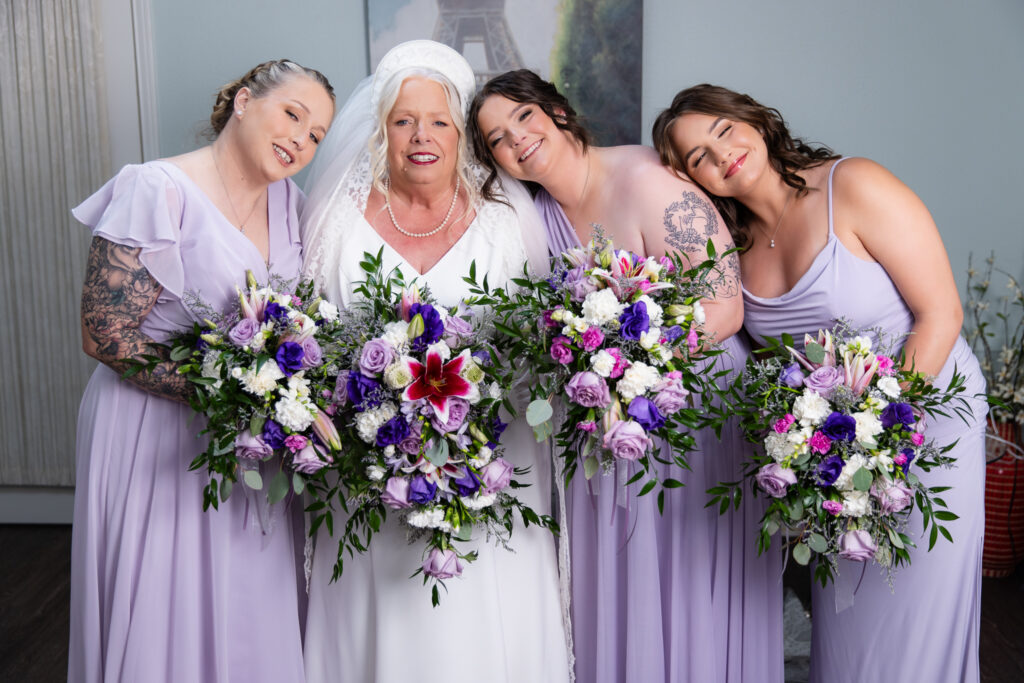 elderly bride smiling with bridemaids holding colorful purple bouquets in bridal suite at The Abbey