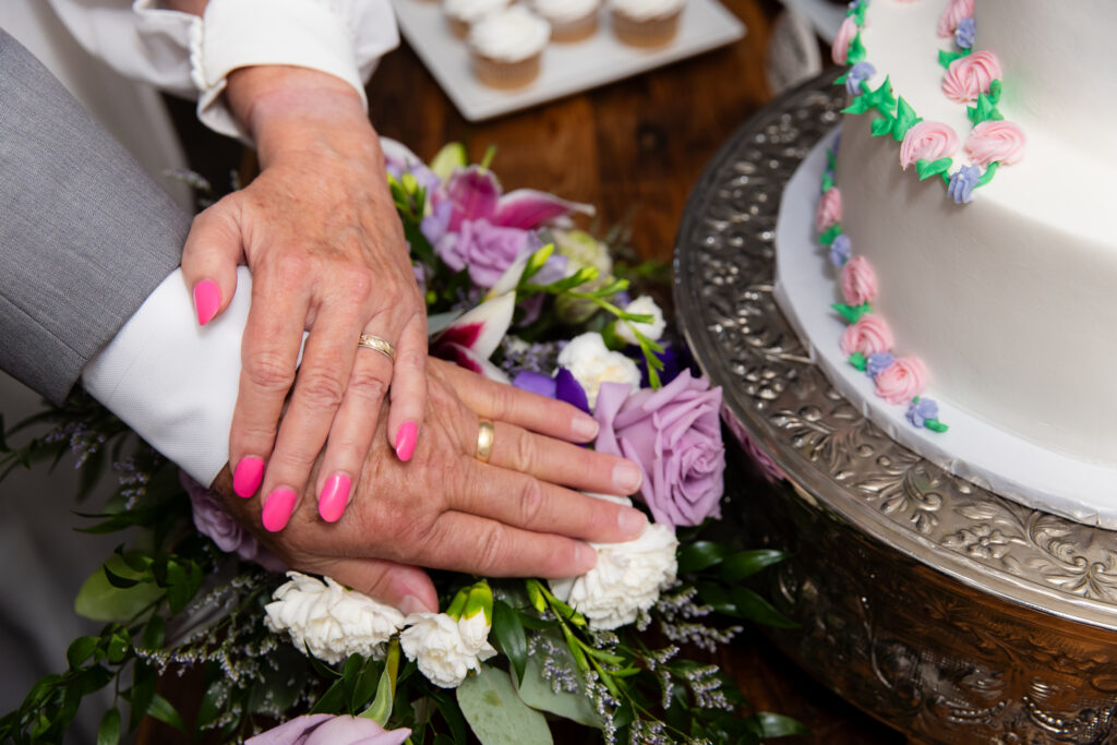 close up of two elderly hands wearing their wedding rings on top of florals next to their wedding cake during reception for their 50th vow renewal celebration