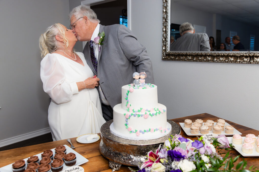 elderly bride and groom kissing behind cute white cake with small pink and green floral decor and a cartoon replica of the couple as a cake toopper