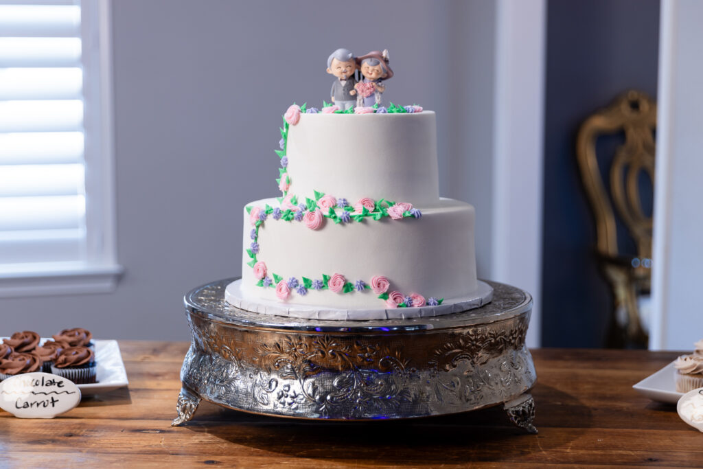 2-tier white cake with simple pink, purple and green floral decor and a cute cartoon cake topper with a happy elderly couple