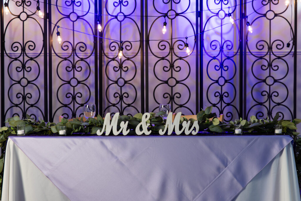 elegant sweetheart table with white linen, mr and mrs sign, greenery and blue uplighting