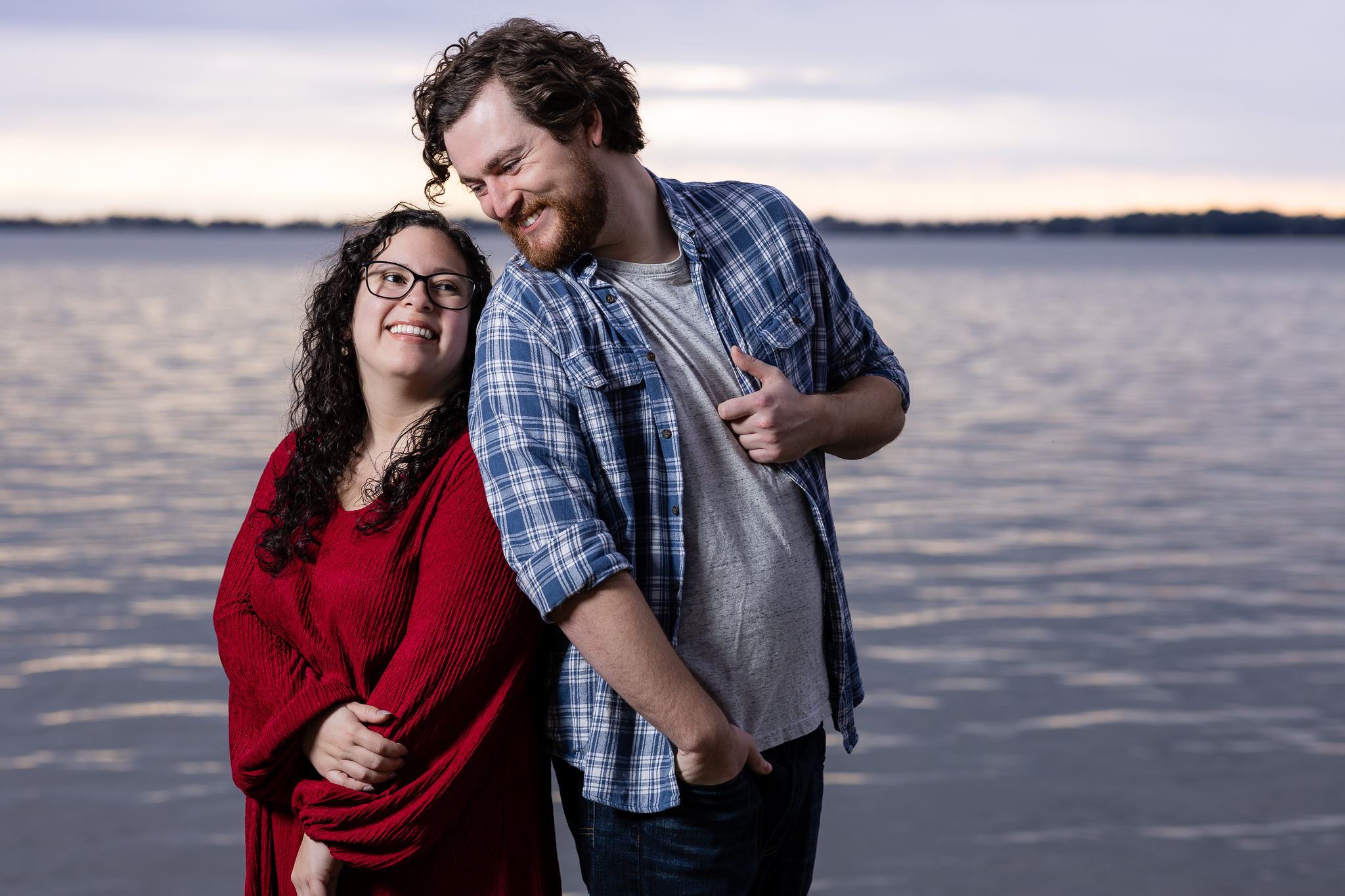 Engaged couple laughing towards each other with lake and sunset behind them during Mabank lakehouse engagement session