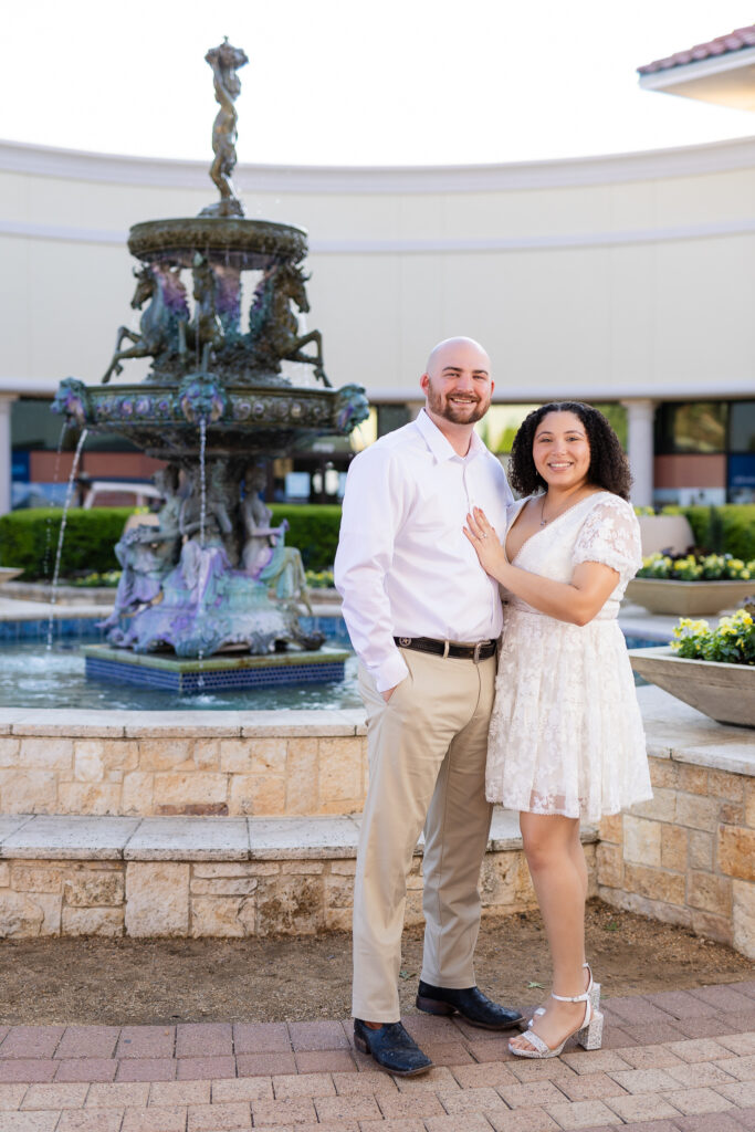 engaged man and woman wearing all white standing together by fountain in Rockwall Texas during engagement session