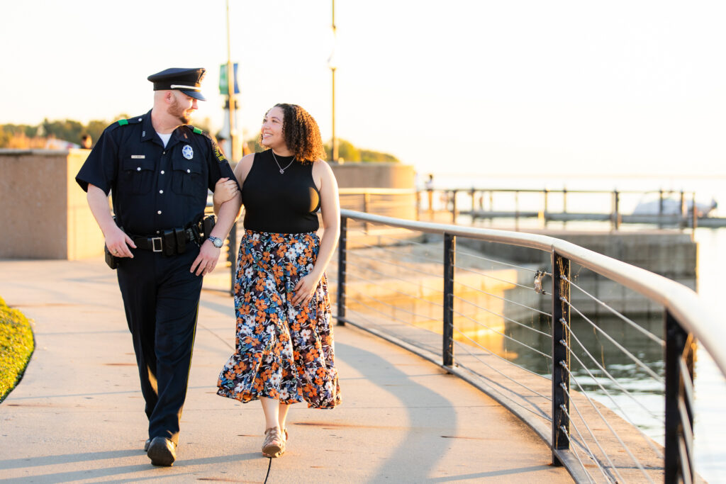 man in police uniform and woman in flowy dress walking along lake in Rockwall during engagement shoot