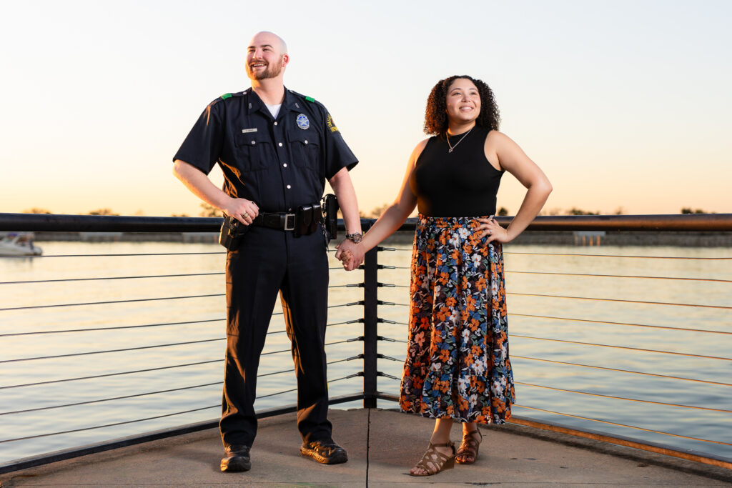 engaged man in police uniform and woman in flowing flower dress holding hands looking out by lake during Rockwall engagement shoot