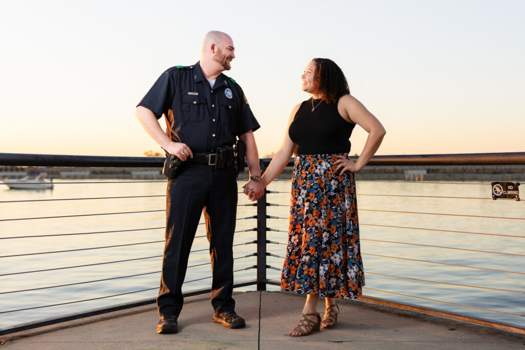 man in police uniform and woman in flowing flower dress holding hands looking at each other by lake during Rockwall engagement shoot